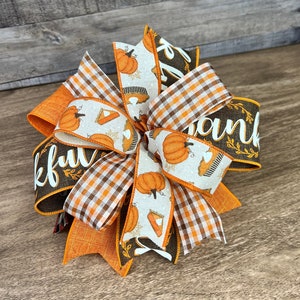 Op de afbeelding: Een Thanksgiving-strik met oranje, bruin en wit geruite en pompoen-gepatroneerde linten. Het lint met het woord "Thankful" is bruin met witte letters en bruine bladeren.