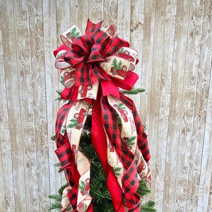 May include: Red and black buffalo plaid Christmas tree topper with a large bow and red and white ribbon with a vintage truck print. The ribbon is curled and tied in a bow at the top.