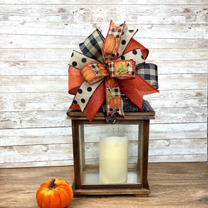 May include: A wooden lantern with a glass panel and a white candle inside. The lantern is decorated with a large, multi-colored bow made of burlap, plaid, and polka dot ribbon. A small orange velvet pumpkin sits on a wooden surface in front of the lantern.