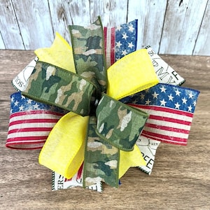 May include: A patriotic bow with red, white, and blue fabric, camouflage fabric, and yellow fabric. The fabric has a distressed look and the bow is tied in a decorative knot.