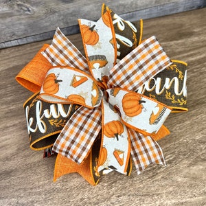 Op de afbeelding: Een Thanksgiving-strik met oranje, bruin en wit lint. Het lint heeft een geruit patroon, een pompoen- en taartontwerp en het woord "Thankful" in bruin schrift.