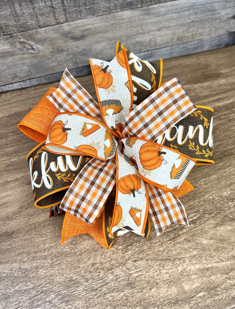 Op de afbeelding: Een Thanksgiving-strik met oranje, bruin en wit lint. Het lint heeft een geruit patroon, een pompoen- en taartontwerp en het woord "Thankful" in bruin schrift.