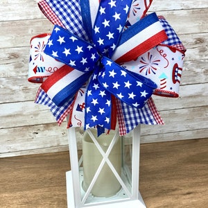 May include: A white lantern with a red, white and blue bow on top. The bow is made of various patterned ribbons, including a blue ribbon with white stars, a red, white and blue striped ribbon, and a white ribbon with red, white and blue fireworks.