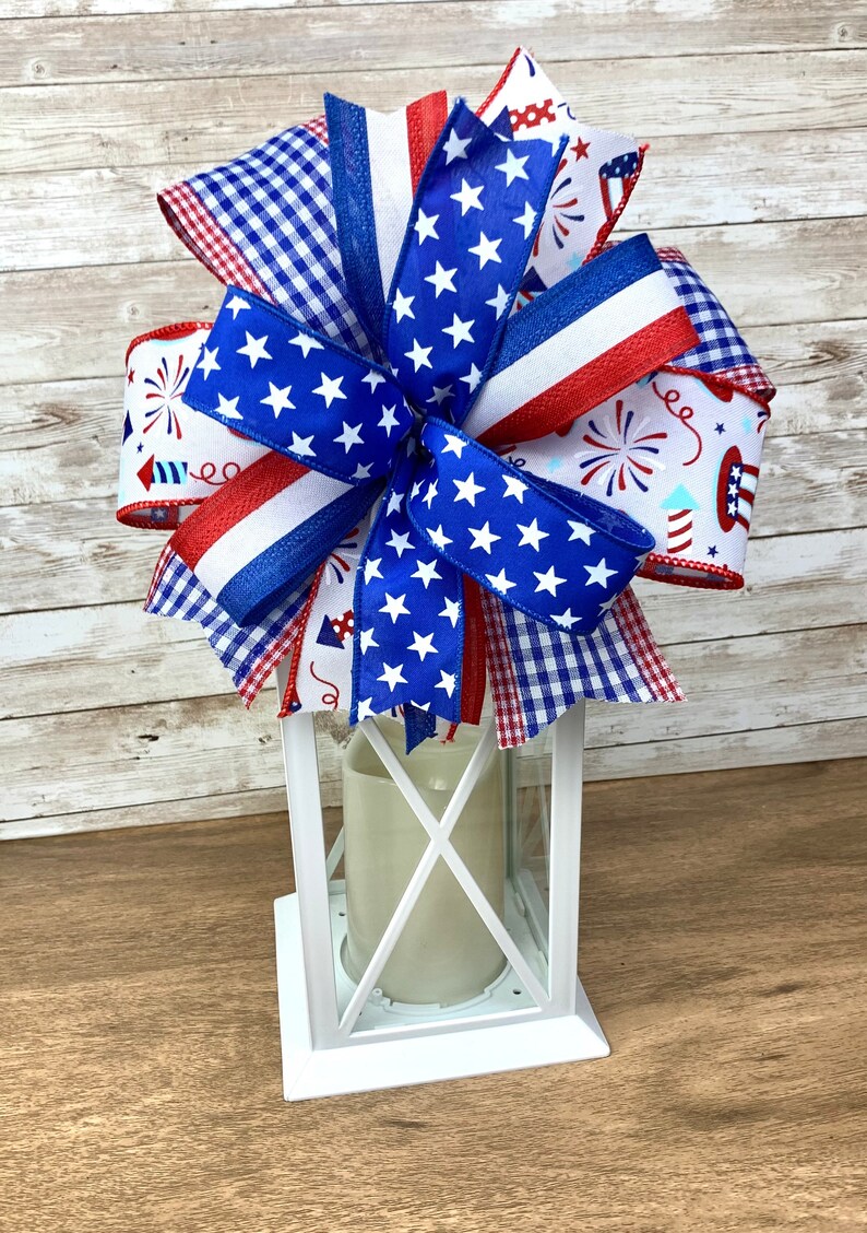 May include: A white lantern with a red, white and blue bow on top. The bow is made of various patterned ribbons, including a blue ribbon with white stars, a red, white and blue striped ribbon, and a white ribbon with red, white and blue fireworks.