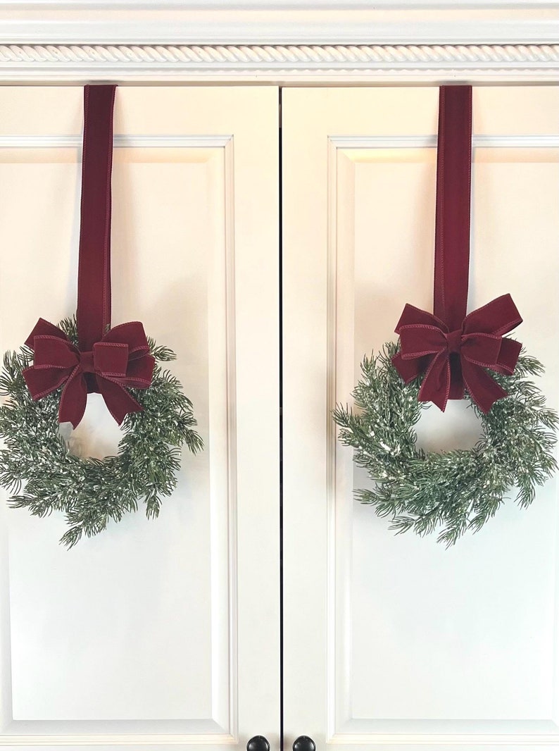 May include: Two Christmas wreaths with faux evergreen branches and burgundy velvet bows hang from white cabinet doors. The wreaths are decorated with a dusting of white, creating a festive holiday display.