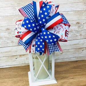 May include: A white lantern with a red, white and blue bow on top. The bow is made of ribbon with stars, stripes and a gingham pattern.