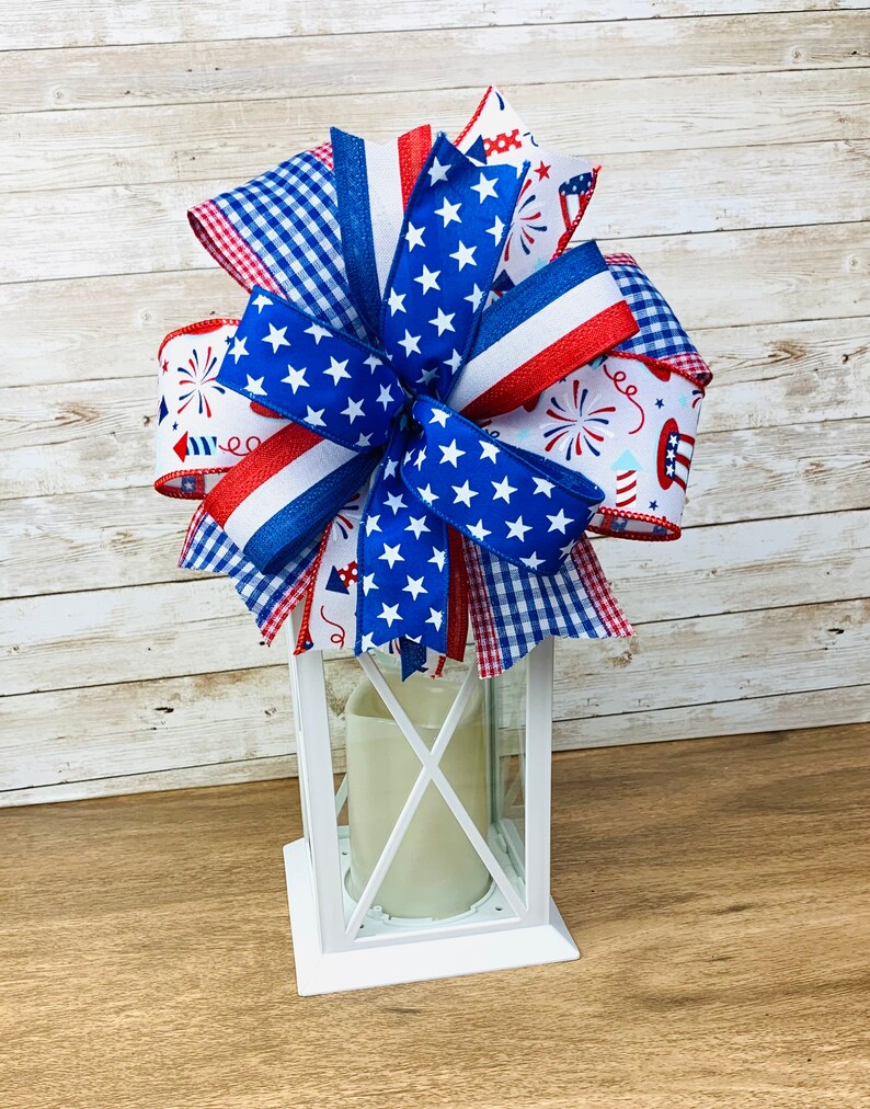 May include: A white lantern with a red, white and blue bow on top. The bow is made of ribbon with stars, stripes and a gingham pattern.