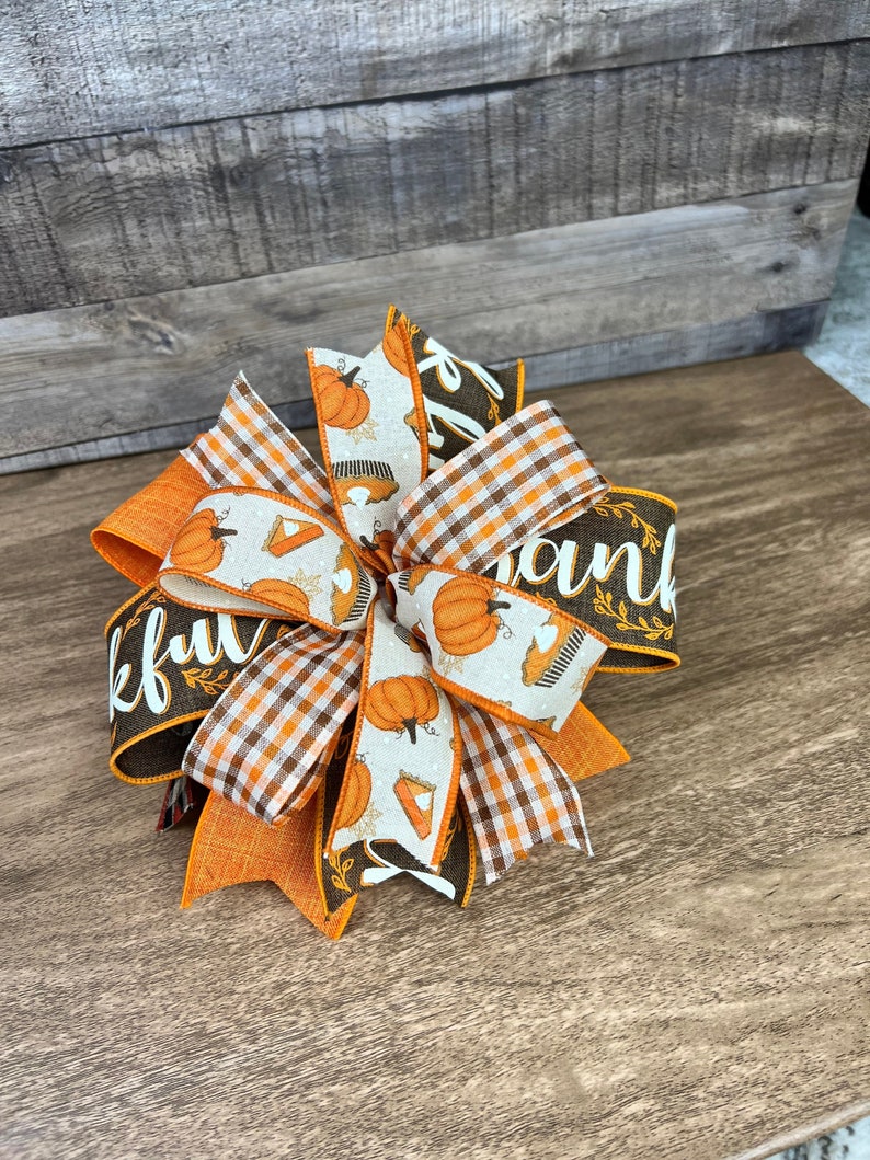 Op de afbeelding: Een Thanksgiving-strik met oranje, bruin en wit geruite en pompoen-gepatroneerde linten. Het lint met het woord "Thankful" is bruin met witte letters en bruine bladeren.