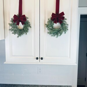 May include: Two white kitchen cabinet doors are adorned with festive holiday decor. Each door features a small evergreen wreath with a burgundy velvet bow, suspended by a matching ribbon. The scene evokes a sense of seasonal cheer.