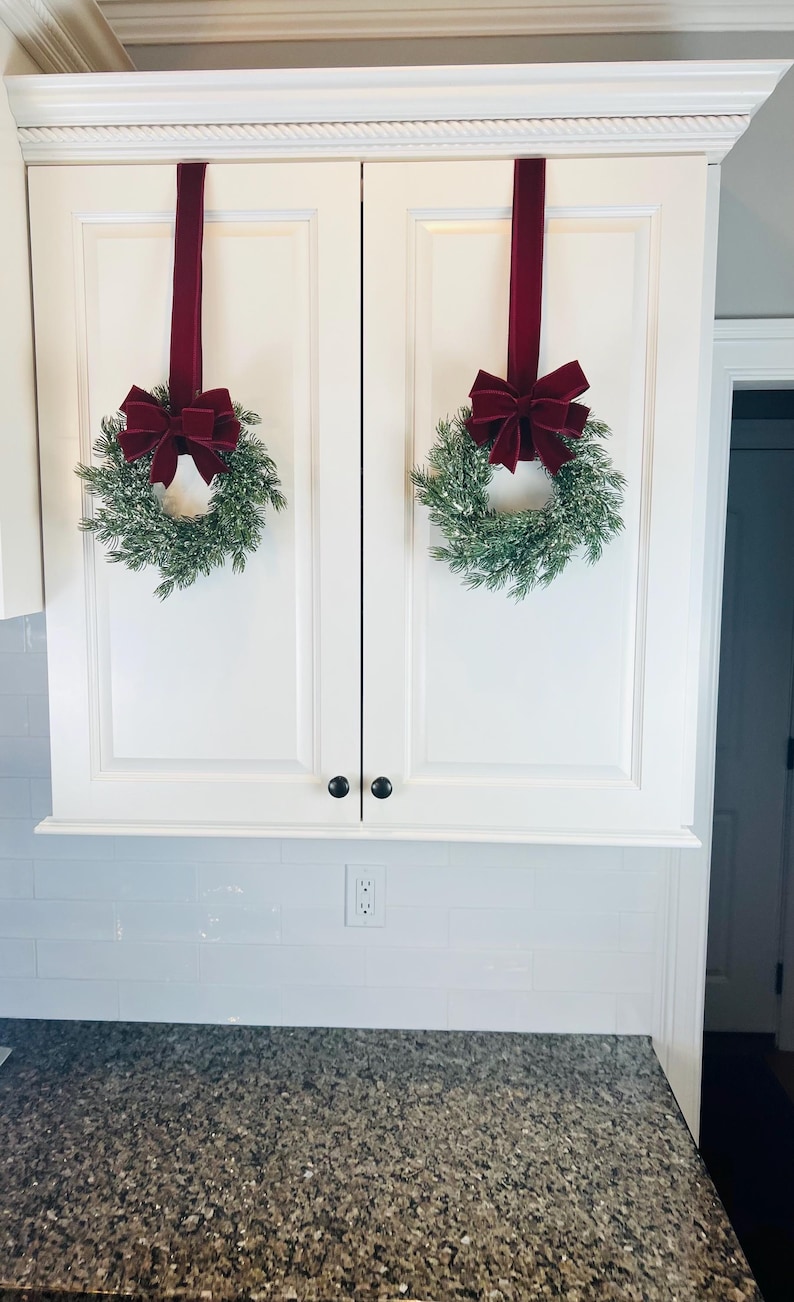 May include: Two white kitchen cabinet doors are adorned with festive holiday decor. Each door features a small evergreen wreath with a burgundy velvet bow, suspended by a matching ribbon. The scene evokes a sense of seasonal cheer.
