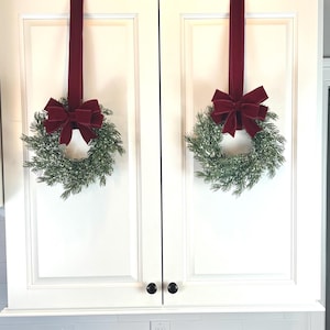 May include: Two Christmas wreaths with red velvet bows hang from a white cabinet. The wreaths are made of green pine branches and are decorated with a dusting of white. The cabinet has black knobs and is above a white tiled backsplash.