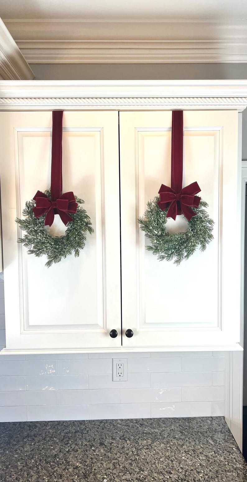 May include: Two Christmas wreaths with red velvet bows hang from a white cabinet. The wreaths are made of green pine branches and are decorated with a dusting of white. The cabinet has black knobs and is above a white tiled backsplash.