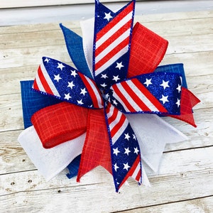 May include: A red, white, and blue patriotic bow with glitter stars and stripes. The bow is made of ribbon and is perfect for decorating for the Fourth of July.
