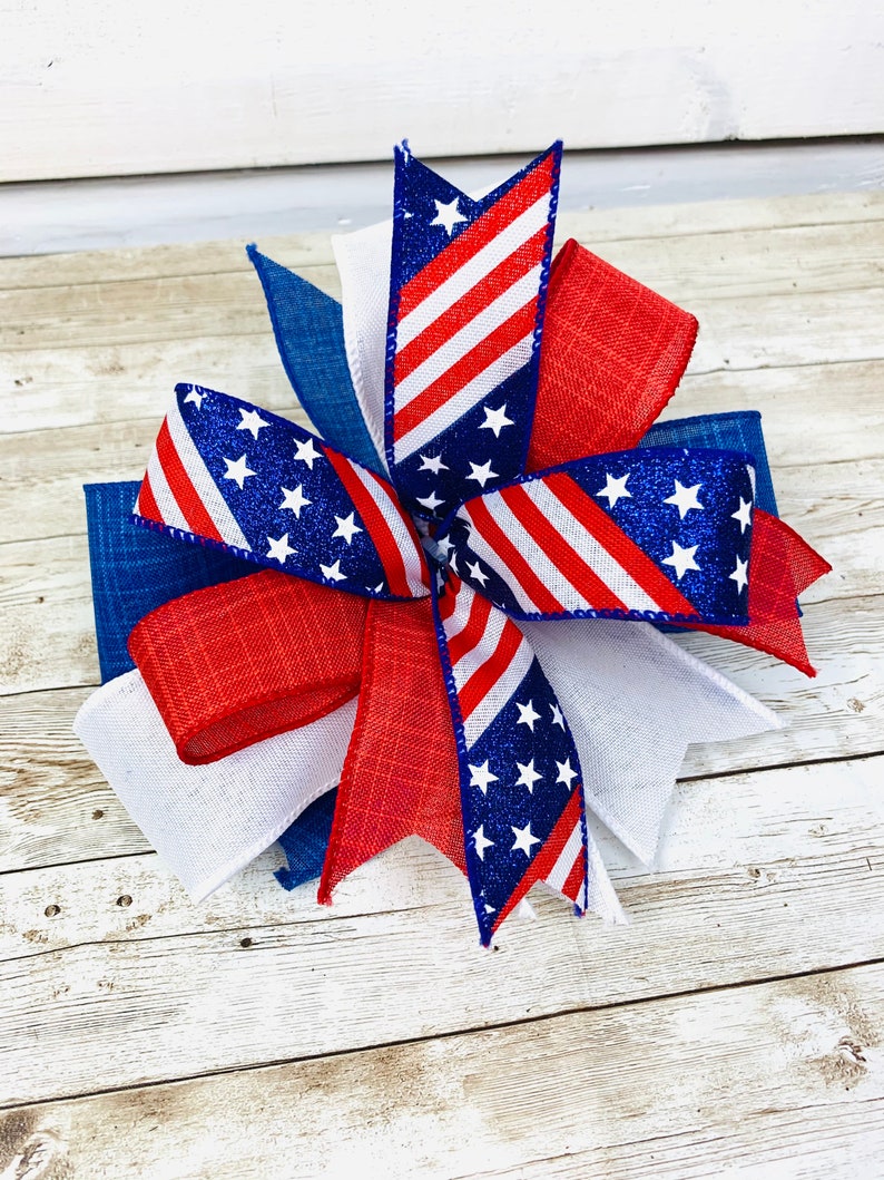 May include: A red, white, and blue patriotic bow with glitter stars and stripes. The bow is made of ribbon and is perfect for decorating for the Fourth of July.
