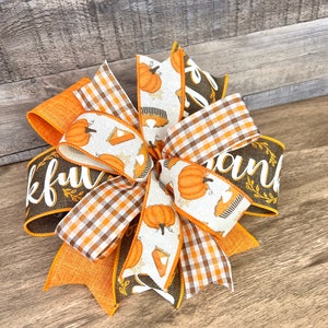 Op de afbeelding: Een bruine en oranje Thanksgiving-thema strik met een geruit patroon en een lint met de tekst "Thankful" en een ander lint met de tekst "Thank"