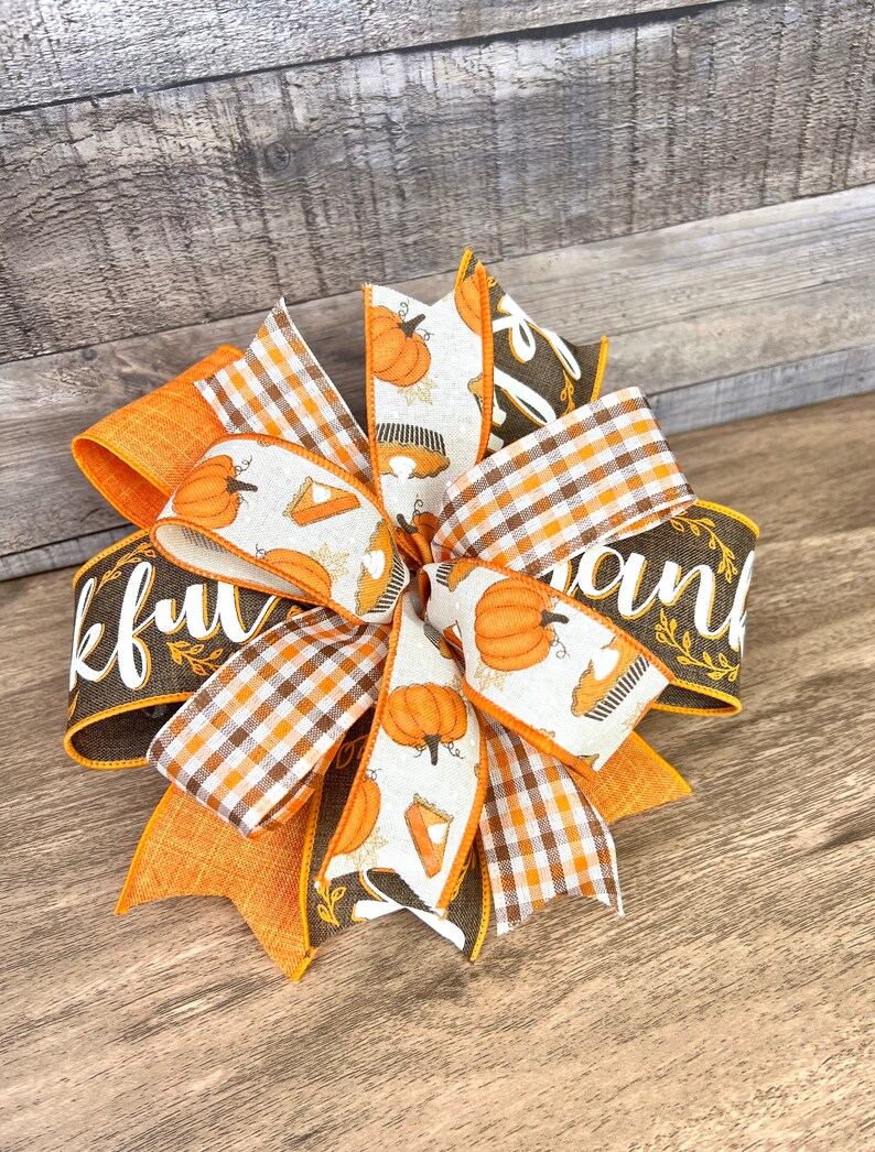 Op de afbeelding: Een bruine en oranje Thanksgiving-thema strik met een geruit patroon en een lint met de tekst "Thankful" en een ander lint met de tekst "Thank"