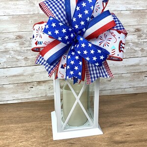 May include: A white lantern with a red, white and blue bow on top. The bow is made of various patterned ribbons, including stars and fireworks.