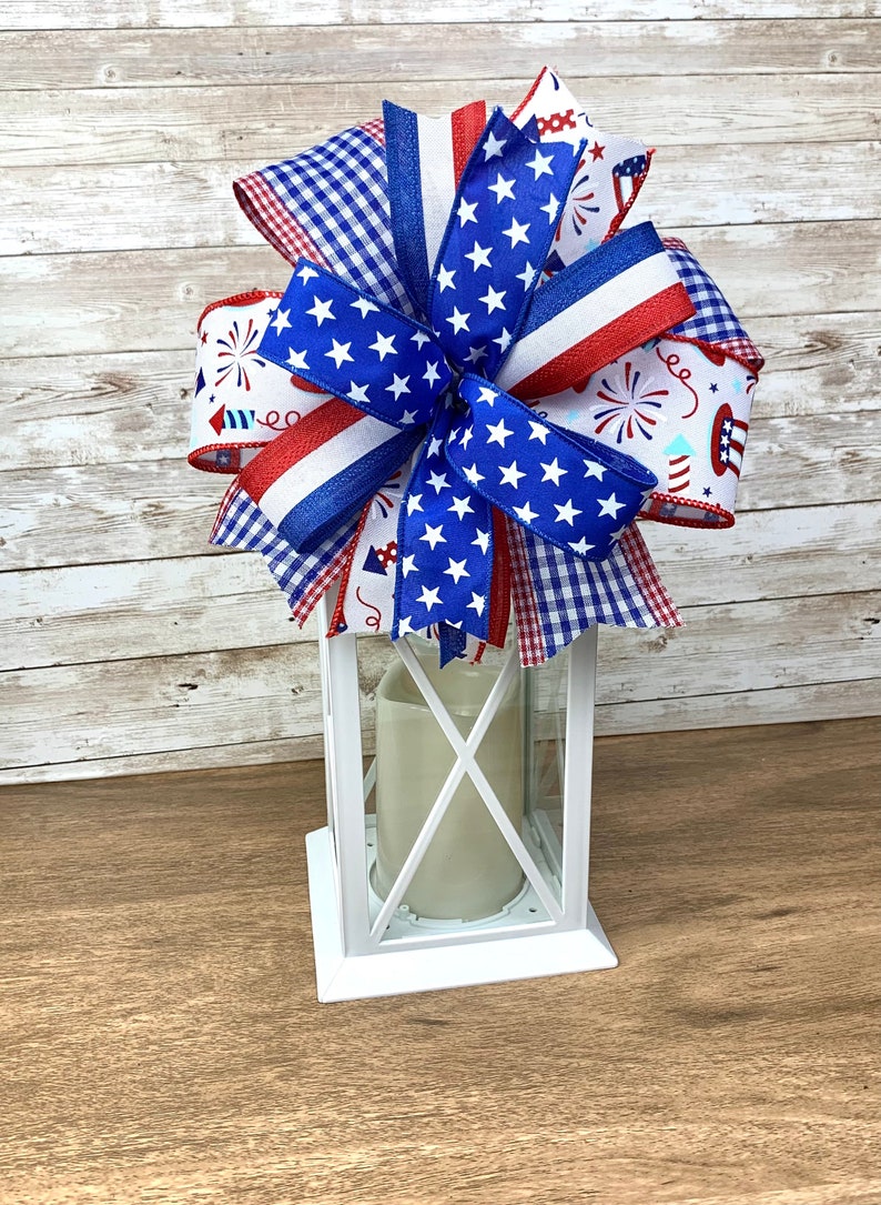 May include: A white lantern with a red, white and blue bow on top. The bow is made of various patterned ribbons, including stars and fireworks.