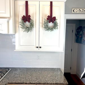 May include: Two white kitchen cabinets with a decorative crown molding. Each cabinet door has a small, round black knob. Two wreaths with green foliage and burgundy bows hang from the cabinets with burgundy ribbons. The countertop is granite.