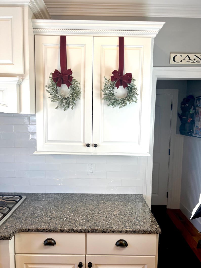 May include: Two white kitchen cabinets with a decorative crown molding. Each cabinet door has a small, round black knob. Two wreaths with green foliage and burgundy bows hang from the cabinets with burgundy ribbons. The countertop is granite.