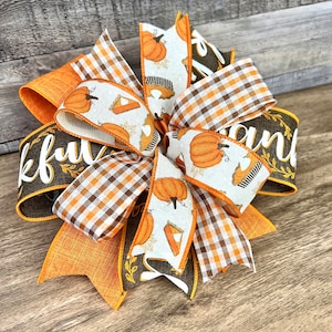 Op de afbeelding: Een Thanksgiving-strik met oranje, bruin en wit lint. Het lint heeft een patroon van pompoenen en taartpunten. De strik is vastgebonden met een bruin lint waarop "Thankful" en "and" staat.