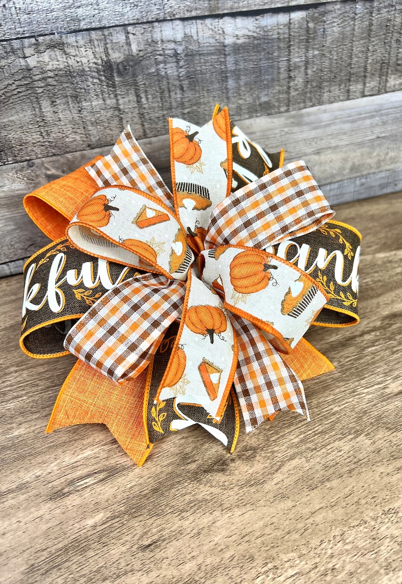Op de afbeelding: Een Thanksgiving-strik met oranje, bruin en wit lint. Het lint heeft een patroon van pompoenen en taartpunten. De strik is vastgebonden met een bruin lint waarop "Thankful" en "and" staat.