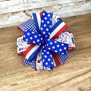 May include: A red, white and blue bow made with patterned ribbon featuring stars, stripes and fireworks. The bow is tied in a knot and has a layered look.