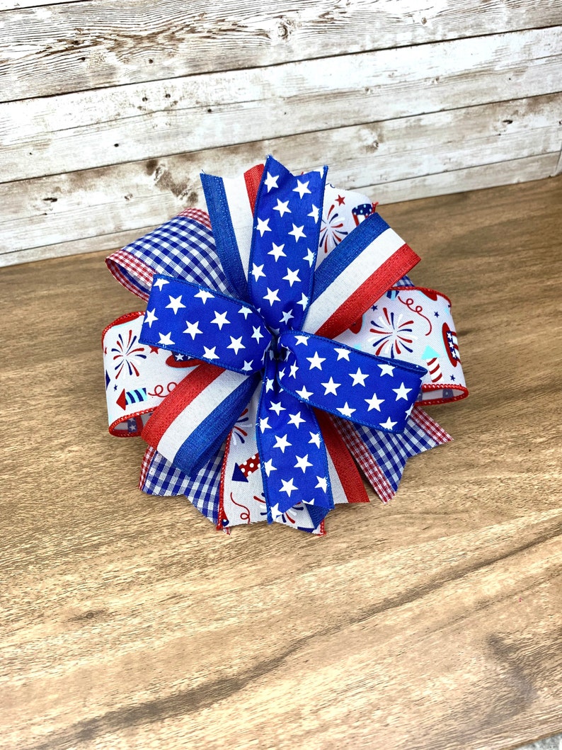 May include: A red, white and blue bow made with patterned ribbon featuring stars, stripes and fireworks. The bow is tied in a knot and has a layered look.