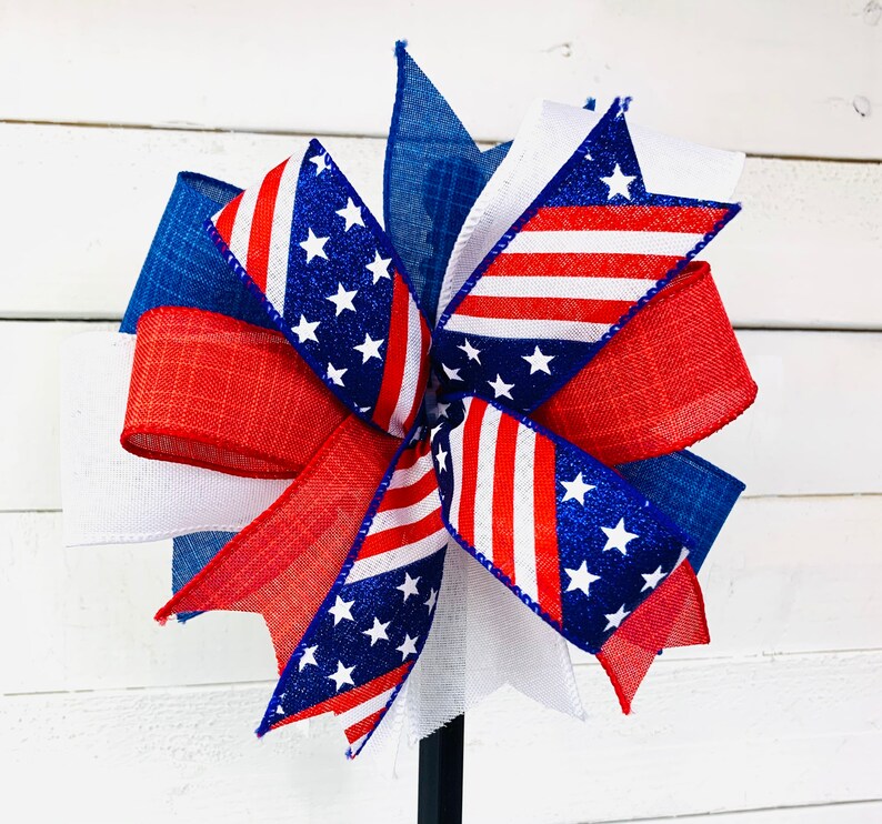 May include: A red, white, and blue patriotic bow with a star-spangled design. The bow is made of fabric and is tied with a ribbon. It is perfect for decorating for the Fourth of July or other patriotic holidays.