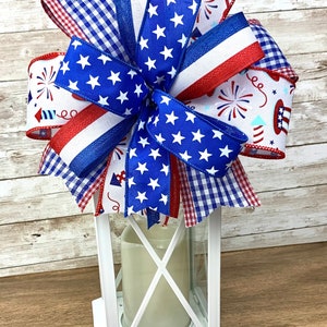 May include: A white lantern with a red, white and blue bow on top. The bow is made of various patterned ribbons, including stars and stripes, gingham and fireworks.