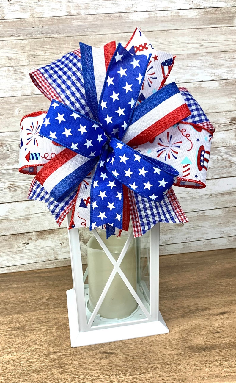 May include: A white lantern with a red, white and blue bow on top. The bow is made of various patterned ribbons, including stars and stripes, gingham and fireworks.