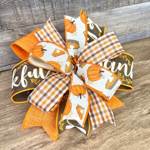 Op de afbeelding: Een Thanksgiving-strik met oranje, bruine en witte linten. De linten hebben een pompoen- en taartpatroon, een ruitpatroon en de woorden "Thankful" en "and".