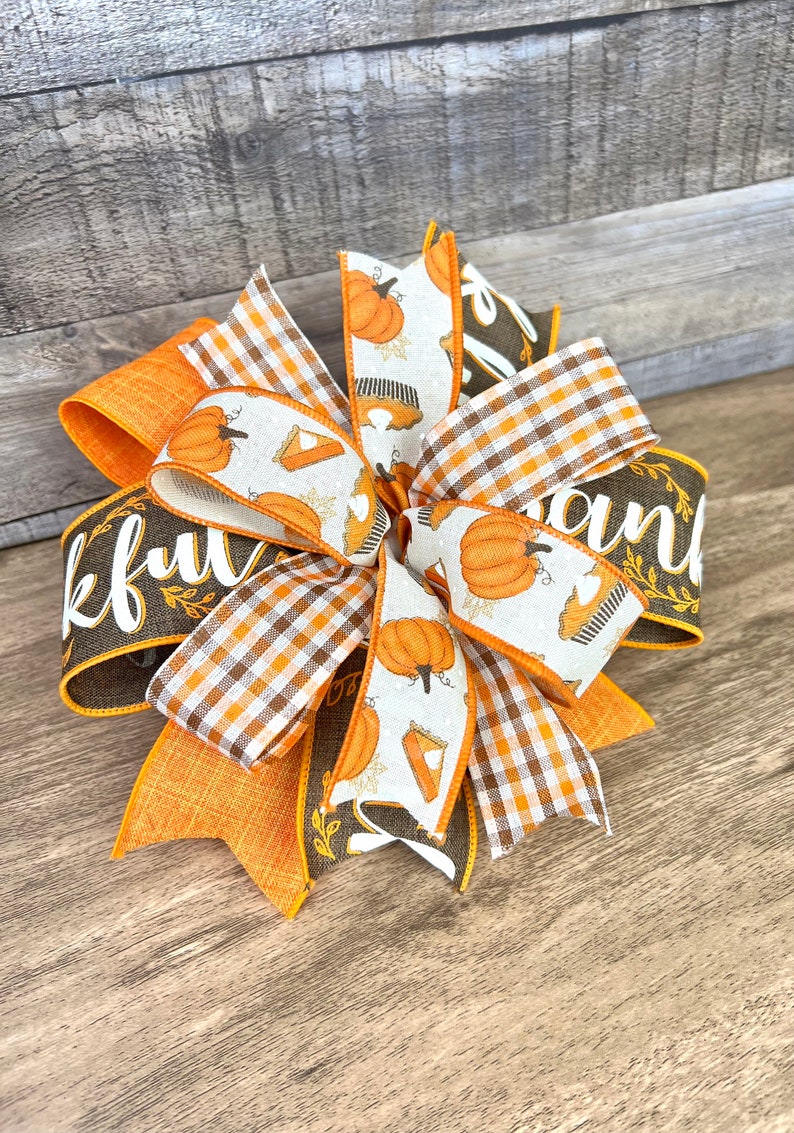 Op de afbeelding: Een Thanksgiving-strik met oranje, bruine en witte linten. De linten hebben een pompoen- en taartpatroon, een ruitpatroon en de woorden "Thankful" en "and".