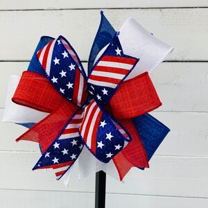 May include: A red, white, and blue bow with a star pattern. The bow is made of ribbon and is tied in a knot. The bow is on a white background.