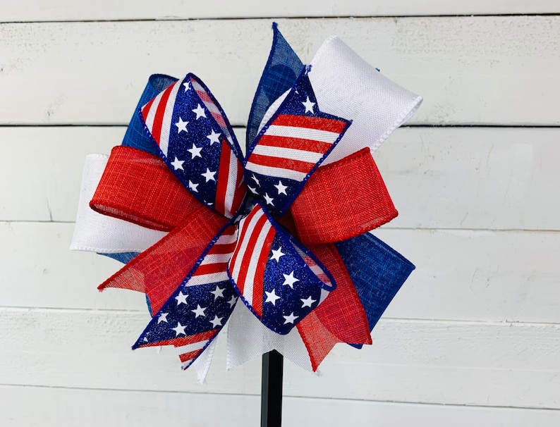 May include: A red, white, and blue bow with a star pattern. The bow is made of ribbon and is tied in a knot. The bow is on a white background.