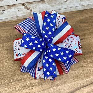 May include: A red, white and blue bow made with various patterned ribbons, including stars, gingham and fireworks. The bow is tied in a knot and has a layered, multi-dimensional look.