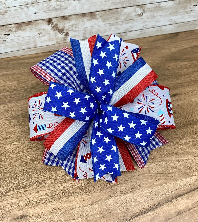 May include: A red, white and blue bow made with various patterned ribbons, including stars, gingham and fireworks. The bow is tied in a knot and has a layered, multi-dimensional look.