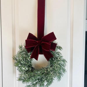 May include: A festive Christmas wreath with faux evergreen branches and a dusting of white snow. A deep burgundy velvet bow and matching ribbon hang the wreath on a white door, creating a classic holiday decoration.