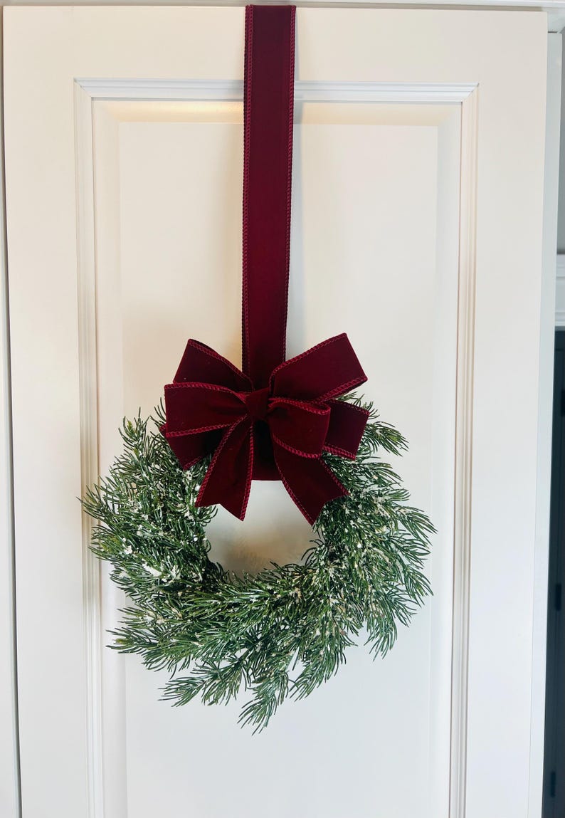May include: A festive Christmas wreath with faux evergreen branches and a dusting of white snow. A deep burgundy velvet bow and matching ribbon hang the wreath on a white door, creating a classic holiday decoration.