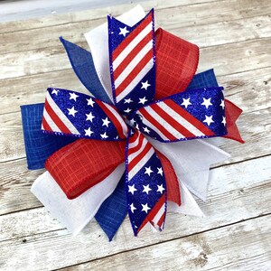 May include: A red, white, and blue bow with glitter stars and stripes. The bow is made of burlap and ribbon.
