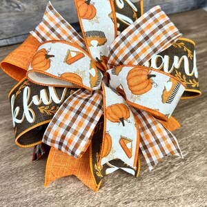 Op de afbeelding: Een Thanksgiving-strik met oranje, bruin en wit geruite en pompoen-gepatroneerde linten. De strik heeft een bruin lint met het woord "Thankful" en een ander bruin lint met het woord "and" erop.