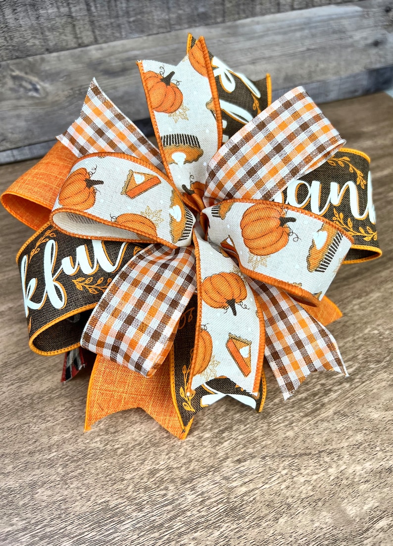 Op de afbeelding: Een Thanksgiving-strik met oranje, bruin en wit geruite en pompoen-gepatroneerde linten. De strik heeft een bruin lint met het woord "Thankful" en een ander bruin lint met het woord "and" erop.