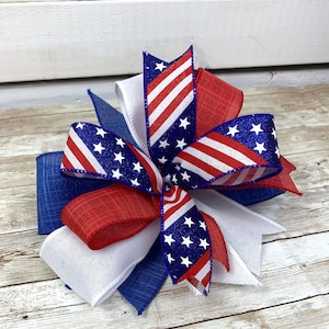 May include: A red, white, and blue bow made of ribbon with a glittery finish. The bow is tied in a knot and has a star pattern.