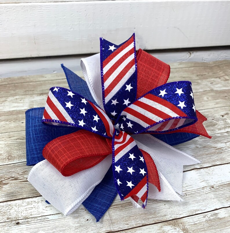 May include: A red, white, and blue bow made of ribbon with a glittery finish. The bow is tied in a knot and has a star pattern.