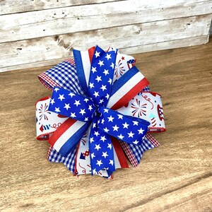 May include: A red, white and blue bow made of ribbon with stars and fireworks patterns. The bow is tied in a knot and is on a wooden surface.