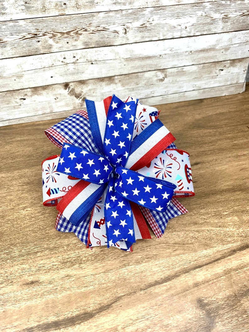 May include: A red, white and blue bow made of ribbon with stars and fireworks patterns. The bow is tied in a knot and is on a wooden surface.