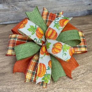 K&ouml;nnte beinhalten: Eine herbstliche Schleife mit orangefarbenem, gr&uuml;nem und wei&szlig;em Band. Das Band hat ein Karomuster, eine einfarbige gr&uuml;ne Farbe und ein wei&szlig;es Band mit einem K&uuml;rbisdruck.
