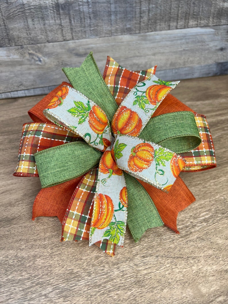 K&ouml;nnte beinhalten: Eine herbstliche Schleife mit orangefarbenem, gr&uuml;nem und wei&szlig;em Band. Das Band hat ein Karomuster, eine einfarbige gr&uuml;ne Farbe und ein wei&szlig;es Band mit einem K&uuml;rbisdruck.