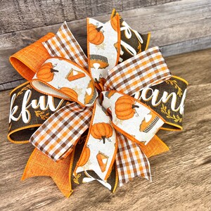 Op de afbeelding: Een bruine, oranje en witte Thanksgiving-strik met een geruit lint en een lint met de woorden "Thankful" en "Thank" erop gedrukt. Het lint heeft ook een patroon van pompoenen en taarten.