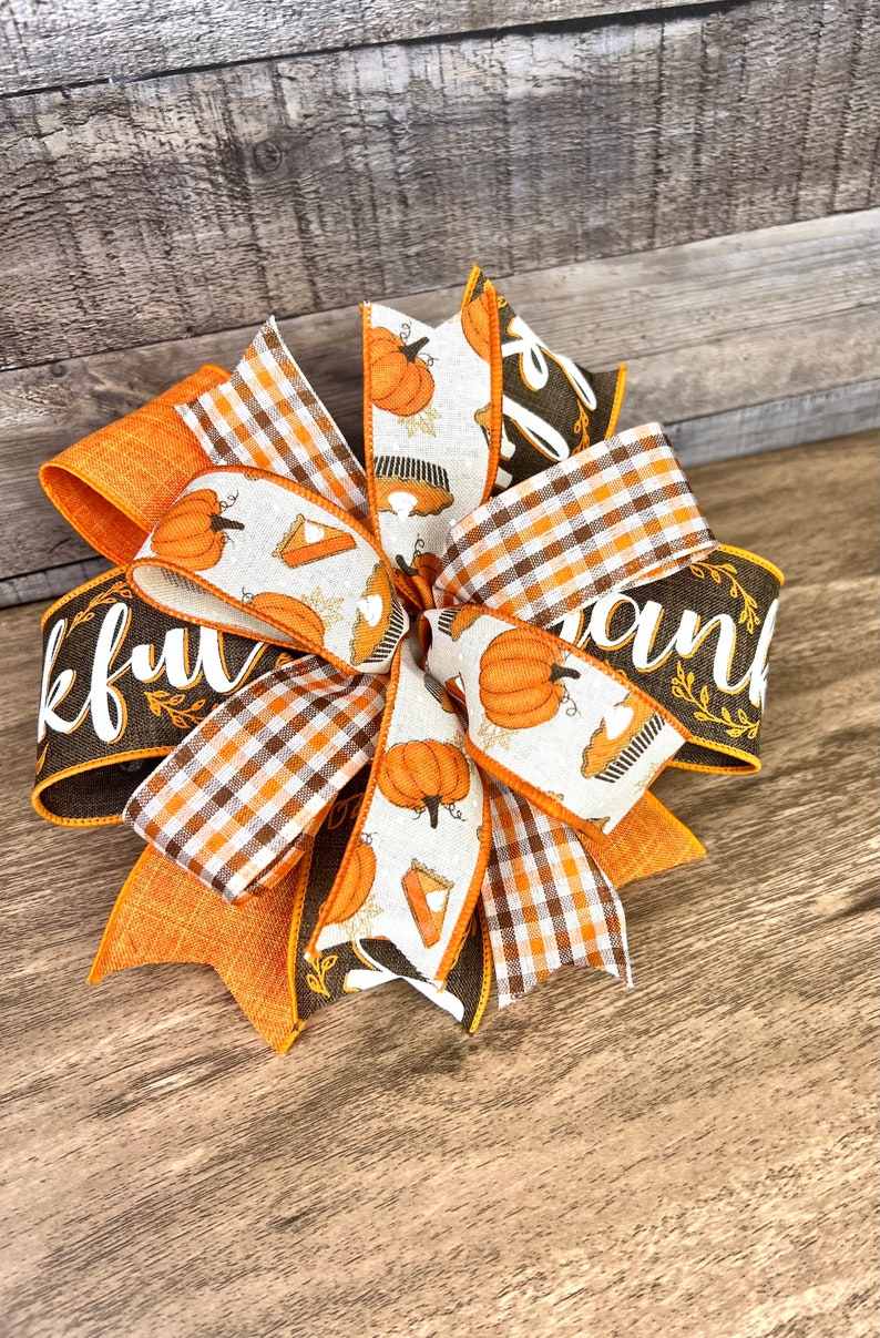 Op de afbeelding: Een bruine, oranje en witte Thanksgiving-strik met een geruit lint en een lint met de woorden "Thankful" en "Thank" erop gedrukt. Het lint heeft ook een patroon van pompoenen en taarten.